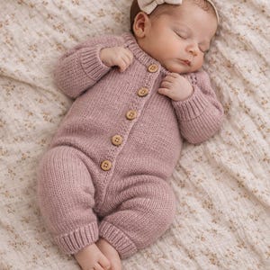 Puede incluir: Un beb&eacute; dormido con un pelele de punto rosa empolvado con botones de madera. El pelele tiene mangas largas y cubre las piernas y los pies del beb&eacute;. Una diadema a juego con lazo completa el conjunto. El beb&eacute; est&aacute; acostado sobre una manta con estampado floral.