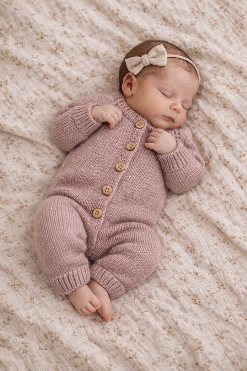 Puede incluir: Un beb&eacute; dormido con un pelele de punto rosa empolvado con botones de madera. El pelele tiene mangas largas y cubre las piernas y los pies del beb&eacute;. Una diadema a juego con lazo completa el conjunto. El beb&eacute; est&aacute; acostado sobre una manta con estampado floral.