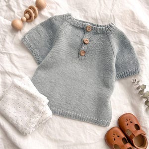 Puede incluir: Una camiseta de bebé de punto azul claro con tres botones de madera, combinada con zapatos de bebé de cuero marrón, un sonajero de madera y una muselina blanca con lunares grises. Los artículos están dispuestos sobre una superficie blanca texturizada.