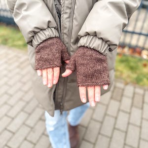 以下が含まれることがあります： 質感のあるニット糸で作られた茶色のフィンガーレスグローブ。手のひらと手の甲を覆い、指先を露出させるシンプルなデザインです。ニット素材には、かすかな霜降り調の風合いが見られます。