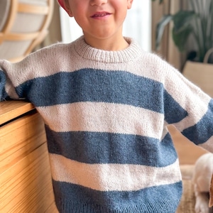 Puede incluir: Un suéter de punto para niño con rayas horizontales en azul claro, blanco roto y azul marino. El suéter tiene cuello redondo y mangas largas. El niño sonríe cerca de un mueble de madera.