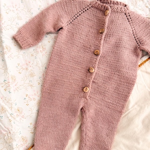 Puede incluir: Un pelele de beb&eacute; tejido de color rosa empolvado, con mangas y perneras largas. El pelele presenta una parte delantera abotonada con botones de madera y pu&ntilde;os y tobillos acanalados. La prenda est&aacute; hecha de un hilo suave y texturizado.