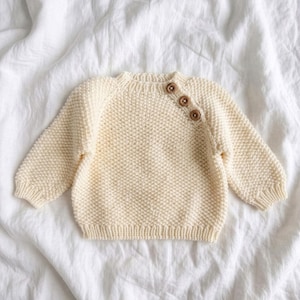 Puede incluir: Un suéter de punto de manga larga en color crema con un patrón texturizado de punto de arroz, cuello redondo acanalado y tres botones de madera marrón colocados en diagonal sobre el hombro izquierdo para un cierre seguro.