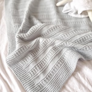 Puede incluir: Una manta de punto gris claro con un patrón texturizado de rayas verticales acanaladas y un borde en punto bobo. Una pequeña muñeca blanca con un detalle amarillo es parcialmente visible en la esquina superior derecha de la tela doblada.