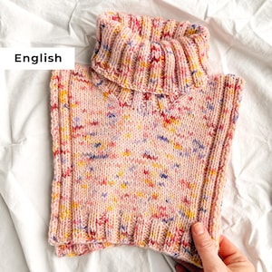 Puede incluir: Un cuello tejido a mano de color rosa con cuello alto acanalado. El cuello está moteado con motas amarillas, azules y rojas. El artículo se muestra sobre una superficie blanca.