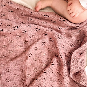 May include: A dusty rose knitted baby blanket featuring a repeating eyelet lace pattern with small, diamond-shaped perforations throughout the fabric. The blanket has a textured border and is draped in soft folds, showcasing the intricate yarn work.