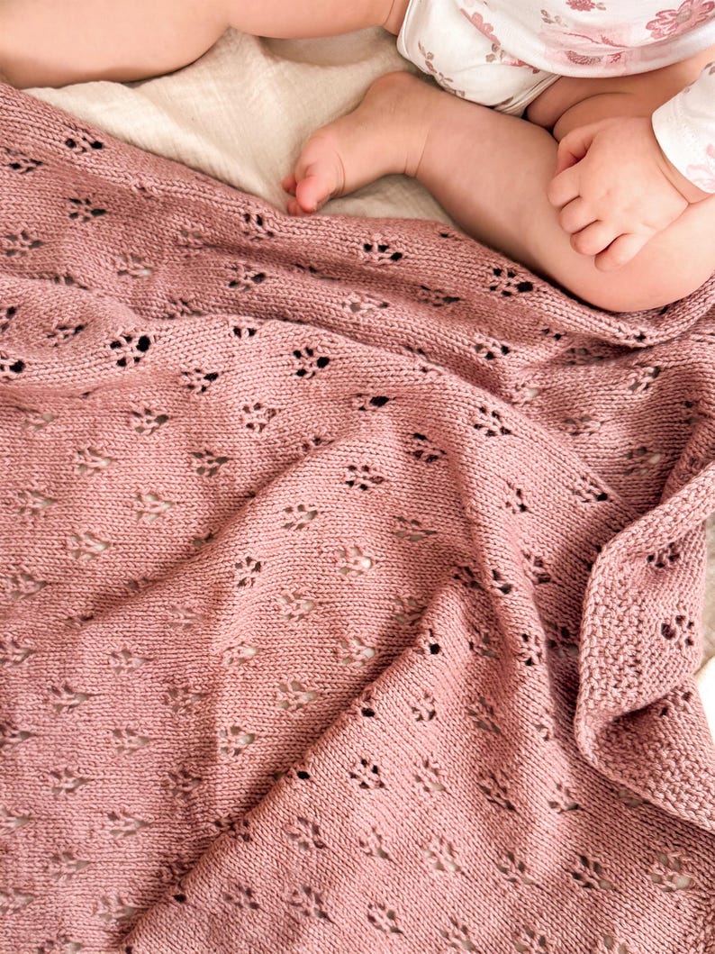 May include: A dusty rose knitted baby blanket featuring a repeating eyelet lace pattern with small, diamond-shaped perforations throughout the fabric. The blanket has a textured border and is draped in soft folds, showcasing the intricate yarn work.