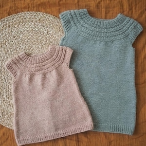 May include: Two knitted sleeveless dresses. One is light pink, the other is a muted blue-green. Both have a detailed, ribbed neckline and a simple, straight silhouette. The dresses are displayed on a textured, neutral-colored surface.