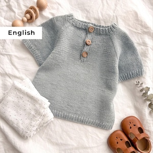 Puede incluir: Una camiseta de bebé de punto azul claro con tres botones de madera. La imagen también muestra zapatos de bebé de cuero marrón, una muselina blanca y gris con lunares y un sonajero de madera. Todos los artículos están colocados sobre un fondo de tela blanca.