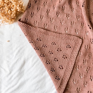 May include: A dusty rose knitted blanket featuring a delicate openwork lace pattern with small repeating floral motifs. The fabric has a textured knit border along the edge, showcasing a soft, consistent yarn construction throughout the piece.