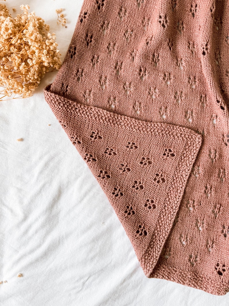 May include: A dusty rose knitted blanket featuring a delicate openwork lace pattern with small repeating floral motifs. The fabric has a textured knit border along the edge, showcasing a soft, consistent yarn construction throughout the piece.