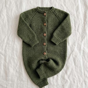 Puede incluir: Un pelele de punto color verde oliva de manga larga con cuello redondo, puños acanalados en muñecas y tobillos, y una tapeta frontal con cinco pequeños botones de madera marrón. La prenda presenta un patrón de punto texturizado con detalle raglán.