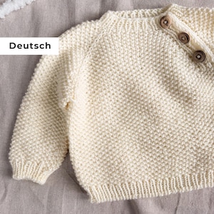 May include: Cream-coloured knitted baby sweater with three wooden buttons on the shoulder. The sweater has a ribbed hem and cuffs. The word "Deutsch" is visible in the image.