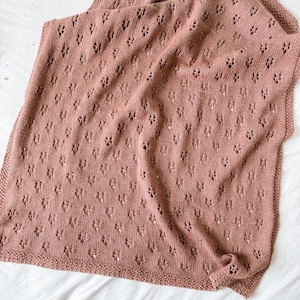 May include: A dusty rose knitted blanket featuring a repeating eyelet lace pattern throughout the fabric. The blanket has a garter stitch border along the edges, creating a textured frame for the delicate, openwork design in the centre.