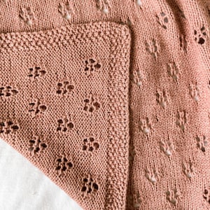 May include: A dusty rose knitted blanket featuring a repeating openwork lace pattern of small flower-like motifs. The fabric has a textured knit border along the edge, showcasing a consistent stitch density and a soft, matte finish throughout.