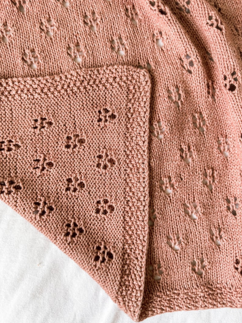 May include: A dusty rose knitted blanket featuring a repeating openwork lace pattern of small flower-like motifs. The fabric has a textured knit border along the edge, showcasing a consistent stitch density and a soft, matte finish throughout.