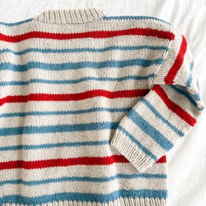 Pode incluir: Um su&eacute;ter de malha com listras horizontais vermelhas e azul claro sobre um fundo bege. O su&eacute;ter tem gola redonda e mangas compridas. O tecido parece ser um tecido de malha macio e texturizado.
