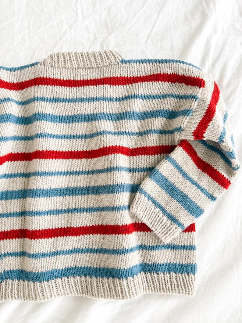 Pode incluir: Um su&eacute;ter de malha com listras horizontais vermelhas e azul claro sobre um fundo bege. O su&eacute;ter tem gola redonda e mangas compridas. O tecido parece ser um tecido de malha macio e texturizado.