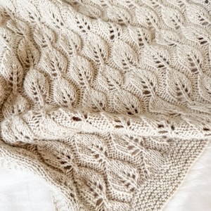 May include: A beige knitted blanket with a leaf pattern. The blanket is folded, showing the intricate design and texture. The leaf pattern is repeated across the surface, creating a cozy and decorative textile.