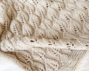 May include: A beige knitted blanket with a leaf pattern. The blanket is folded, showing the intricate design and texture. The leaf pattern is repeated across the surface, creating a cozy and decorative textile.