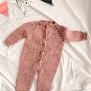 Puede incluir: Un pelele de beb&eacute; de punto color rosa empolvado con mangas largas y botones de madera. La prenda de una pieza se abrocha por delante y tiene pu&ntilde;os y tobillos acanalados. Una pancarta decorativa con banderas rosas, negras y blancas est&aacute; en el fondo.
