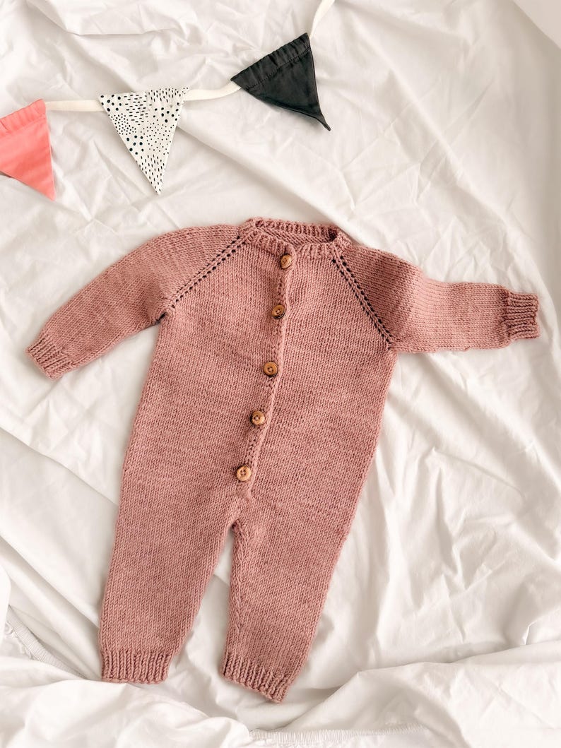 Puede incluir: Un pelele de beb&eacute; de punto color rosa empolvado con mangas largas y botones de madera. La prenda de una pieza se abrocha por delante y tiene pu&ntilde;os y tobillos acanalados. Una pancarta decorativa con banderas rosas, negras y blancas est&aacute; en el fondo.