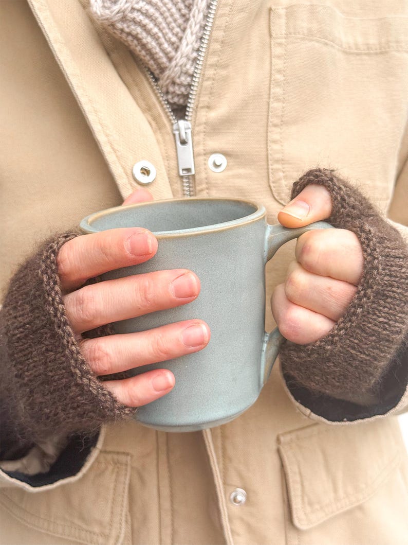 以下が含まれることがあります： 滑らかな仕上げとシンプルな持ち手が付いた水色のセラミック製マグカップ。ダークブラウンのニット製フィンガーレスグローブを着用した手で持たれています。マグカップは縁に繊細なディテールがあり、マットな質感の円筒形のボディをしています。