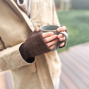 以下が含まれることがあります： ダークブラウンのニット製フィンガーレスグローブを着用した両手が、無地のライトグレーのセラミック製マグカップを持っています。グローブはシンプルなメリヤス編みで、手首と手のひらを覆い、指先は露出したデザインになっています。
