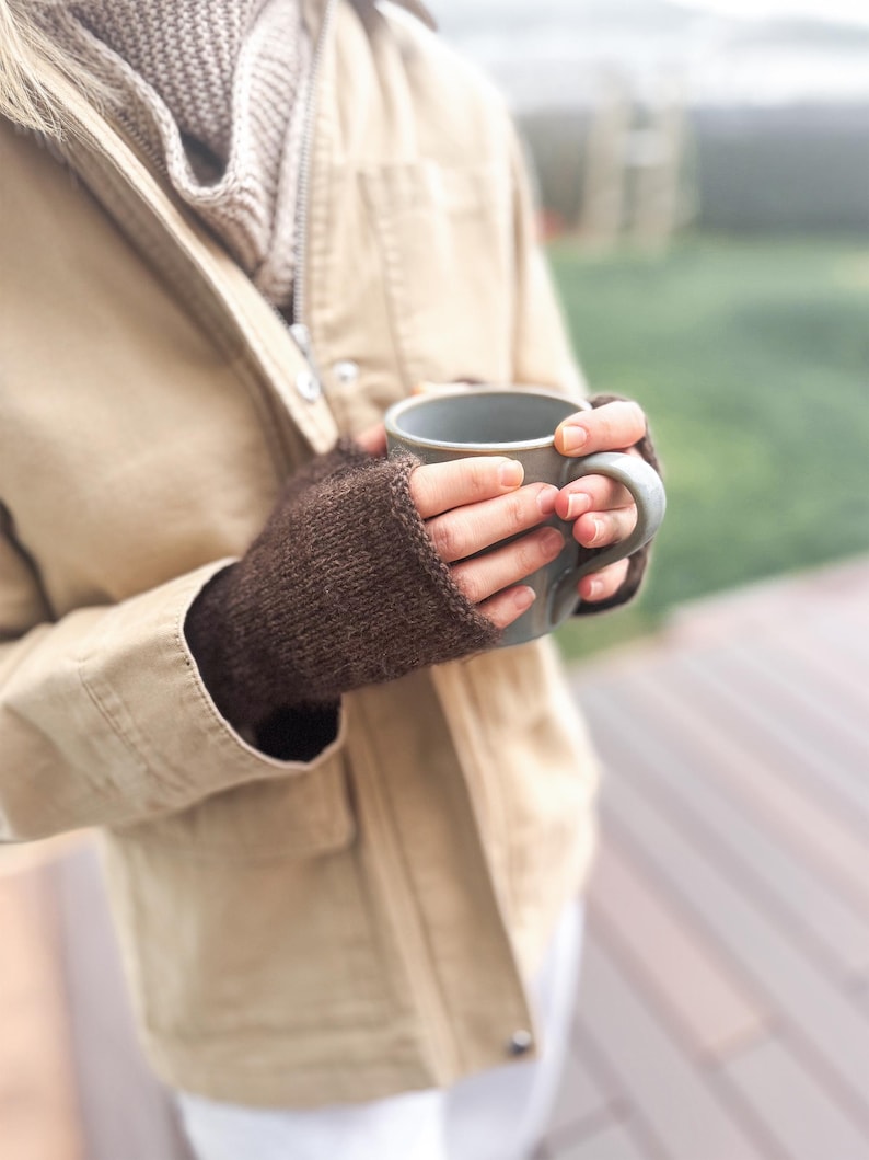 以下が含まれることがあります： ダークブラウンのニット製フィンガーレスグローブを着用した両手が、無地のライトグレーのセラミック製マグカップを持っています。グローブはシンプルなメリヤス編みで、手首と手のひらを覆い、指先は露出したデザインになっています。