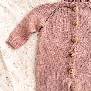 Puede incluir: Mono de beb&eacute; de punto rosa empolvado con mangas largas y botones de madera. El mono est&aacute; hecho de un hilo suave y texturizado y presenta un cuello redondo y pu&ntilde;os acanalados. Se muestra sobre una tela con estampado floral.