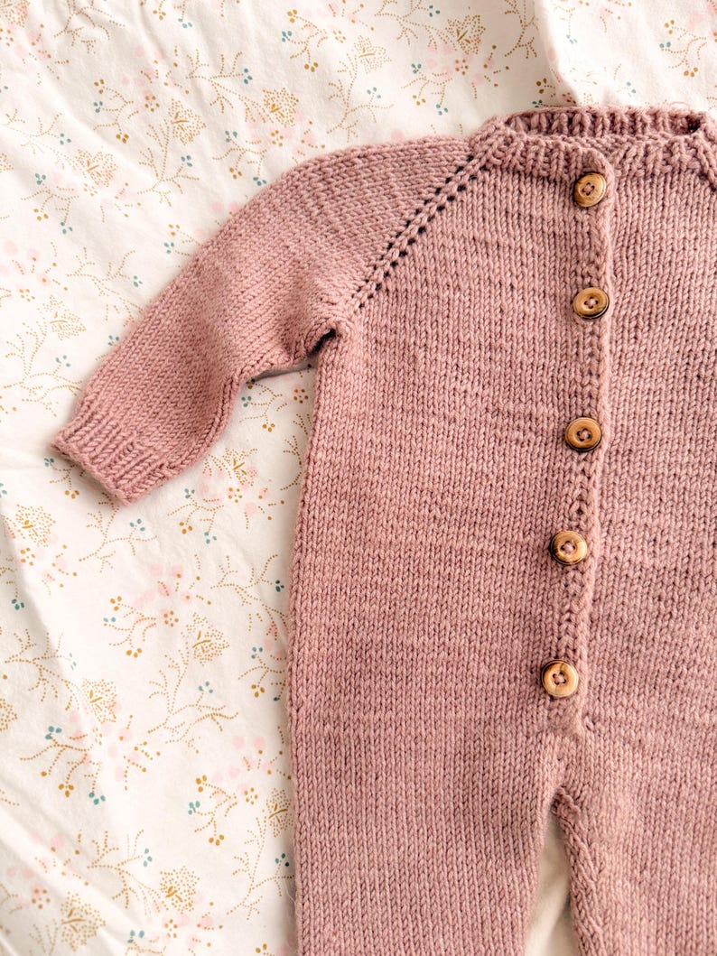 Puede incluir: Mono de beb&eacute; de punto rosa empolvado con mangas largas y botones de madera. El mono est&aacute; hecho de un hilo suave y texturizado y presenta un cuello redondo y pu&ntilde;os acanalados. Se muestra sobre una tela con estampado floral.