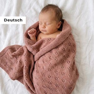Puede incluir: Un bebé dormido envuelto en una manta de punto rosa empolvado. La manta presenta un delicado estampado de hojas y está sobre el bebé en una sábana blanca. La palabra "Deutsch" está en un rectángulo blanco.