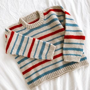 Puede incluir: Un suéter de punto beige con rayas horizontales rojas y azules. El suéter tiene cuello redondo y mangas largas. Las rayas están espaciadas uniformemente en el cuerpo y las mangas.