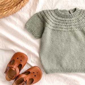 Puede incluir: Un suéter de punto color verde salvia de manga corta con un canesú texturizado y detalles acanalados en el cuello, los puños y el dobladillo, junto a un par de zapatos de cuero marrón con tira en T, aberturas y hebillas metálicas laterales.