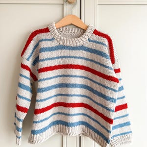 Pode incluir: Um su&eacute;ter de malha com listras horizontais em azul claro, vermelho e branco sujo. O su&eacute;ter est&aacute; pendurado em um cabide de madeira. As mangas e os ombros t&ecirc;m listras vermelhas, e o corpo tem listras azuis e vermelhas alternadas.