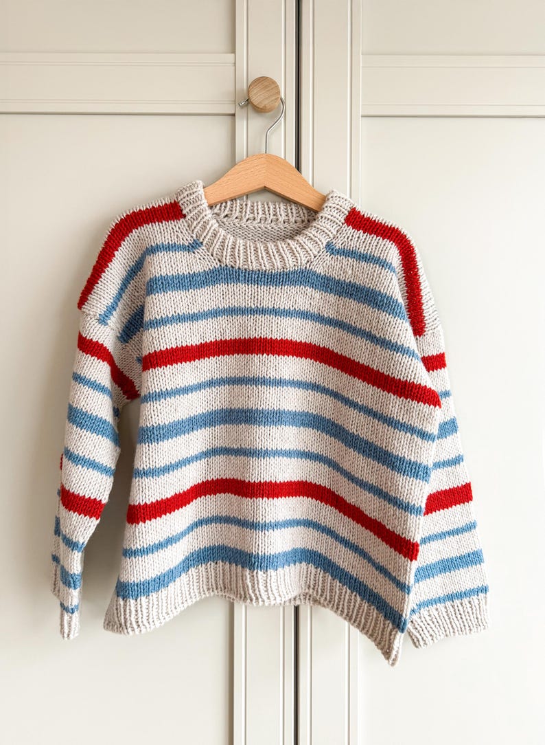 Pode incluir: Um su&eacute;ter de malha com listras horizontais em azul claro, vermelho e branco sujo. O su&eacute;ter est&aacute; pendurado em um cabide de madeira. As mangas e os ombros t&ecirc;m listras vermelhas, e o corpo tem listras azuis e vermelhas alternadas.