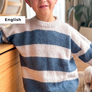 Puede incluir: Un suéter de punto con rayas horizontales en azul claro, blanco roto y azul marino. El suéter tiene cuello redondo y mangas largas. La palabra "English" se muestra en un rectángulo blanco en la imagen.