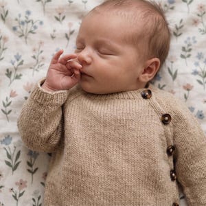 Puede incluir: Un suéter de punto beige de manga larga con cierre lateral de cuatro botones jaspeados de color marrón oscuro. El suéter tiene cuello redondo y textura acanalada, y lo lleva puesto un bebé dormido sobre una tela blanca con un pequeño estampado floral.