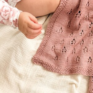 May include: A dusty rose knitted blanket featuring a delicate openwork lace pattern and a textured garter stitch border. The fabric displays a consistent knit structure with small, repeating diamond-shaped eyelet details throughout the textile surface.