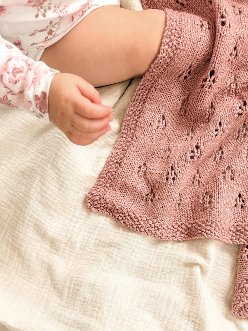 May include: A dusty rose knitted blanket featuring a delicate openwork lace pattern and a textured garter stitch border. The fabric displays a consistent knit structure with small, repeating diamond-shaped eyelet details throughout the textile surface.