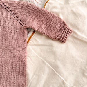 Puede incluir: Primer plano de un su&eacute;ter de punto color rosa empolvado. Se ve la manga y parte del cuerpo, mostrando el patr&oacute;n de punto texturizado. El pu&ntilde;o y el cuello tienen un dise&ntilde;o acanalado. El su&eacute;ter est&aacute; sobre una superficie blanca y con estampado floral.