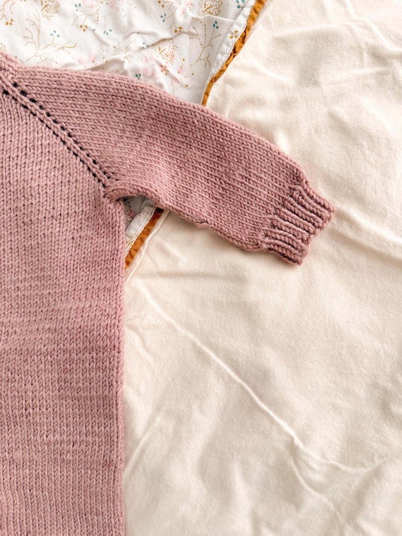 Puede incluir: Primer plano de un su&eacute;ter de punto color rosa empolvado. Se ve la manga y parte del cuerpo, mostrando el patr&oacute;n de punto texturizado. El pu&ntilde;o y el cuello tienen un dise&ntilde;o acanalado. El su&eacute;ter est&aacute; sobre una superficie blanca y con estampado floral.