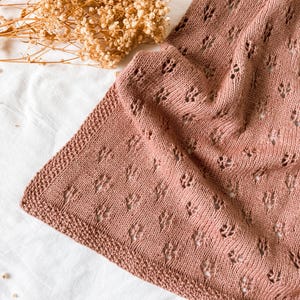 Puede incluir: Una manta de punto color rosa empolvado con un patrón repetitivo de encaje de ojales en toda la tela. La manta tiene un borde texturizado en punto bobo a lo largo de los lados, mostrando un color suave y un diseño calado delicado.