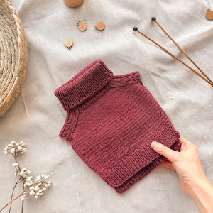 May include: A sleeveless, deep burgundy knit sweater vest featuring a ribbed turtleneck collar and a matching ribbed hem. The garment is crafted with a consistent stockinette stitch pattern throughout the body, showcasing a clean, structured design.