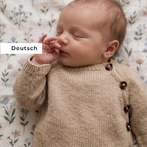 Puede incluir: Un bebé dormido con un suéter de punto beige con botones marrones grandes. El suéter tiene cuello redondo y mangas largas. El bebé está acostado sobre una manta con estampado floral. La palabra "Deutsch" está en un rectángulo blanco en la imagen.