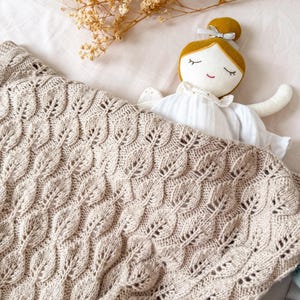 Könnte beinhalten: Eine beige Strickdecke mit Blättermuster, drapiert auf einem Bett. Eine handgefertigte Puppe mit gelbem Dutt und weißem Kleid liegt unter der Decke. Getrocknete Blumen liegen auf dem Bett.
