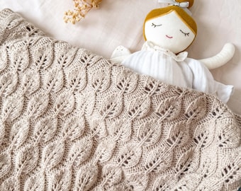 Op de afbeelding: Een beige gebreide deken met een bladerenpatroon, gedrapeerd over een bed. Een handgemaakte pop met een gele knot en een witte jurk ligt onder de deken. Gedroogde bloemen liggen op het bed.