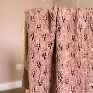 May include: A dusty rose knitted blanket featuring a repeating openwork lace pattern with small, diamond-shaped eyelet holes throughout the fabric. The textile has a soft, textured appearance with a consistent knit stitch and a finished edge.