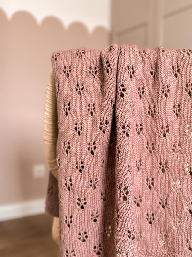 May include: A dusty rose knitted blanket featuring a repeating openwork lace pattern with small, diamond-shaped eyelet holes throughout the fabric. The textile has a soft, textured appearance with a consistent knit stitch and a finished edge.