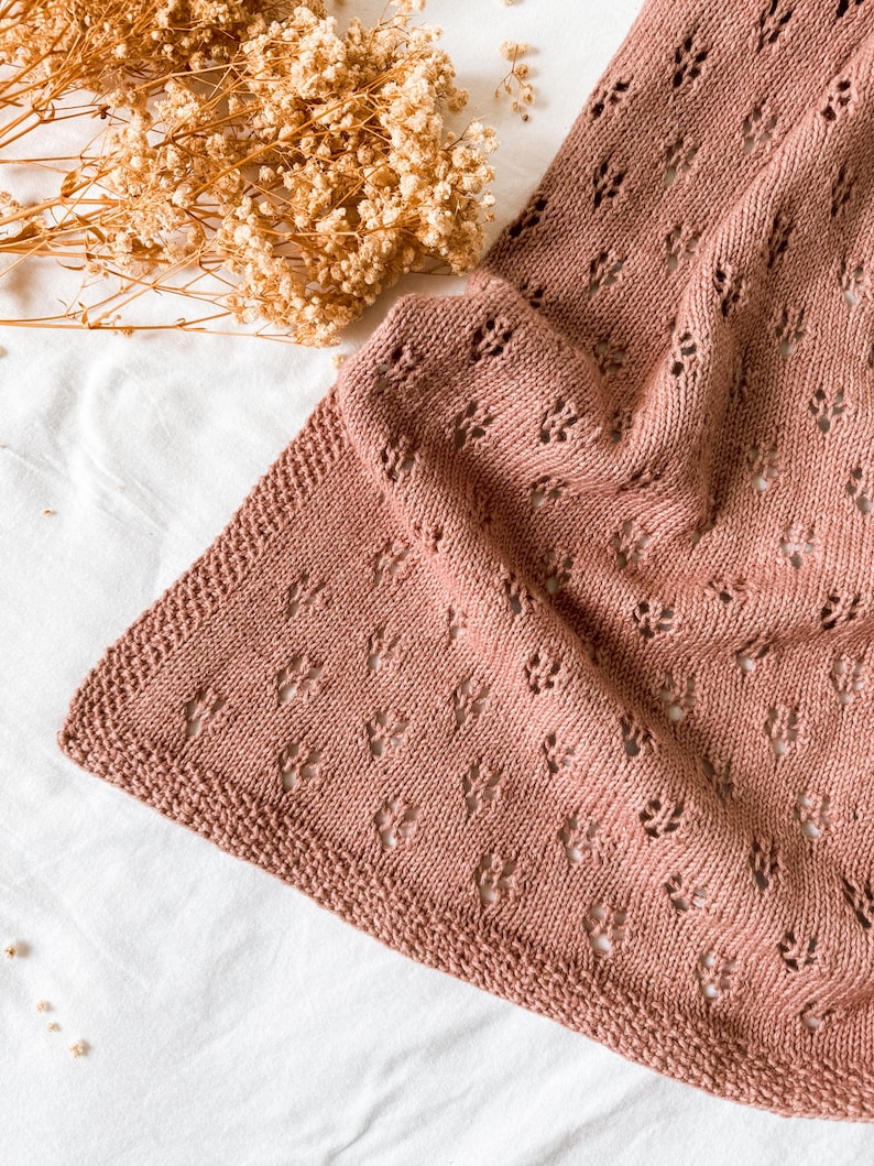May include: A dusty rose knitted blanket featuring a repeating eyelet lace pattern throughout the fabric. The blanket has a textured garter stitch border along the edges, showcasing a soft, uniform colour and a delicate, openwork design.
